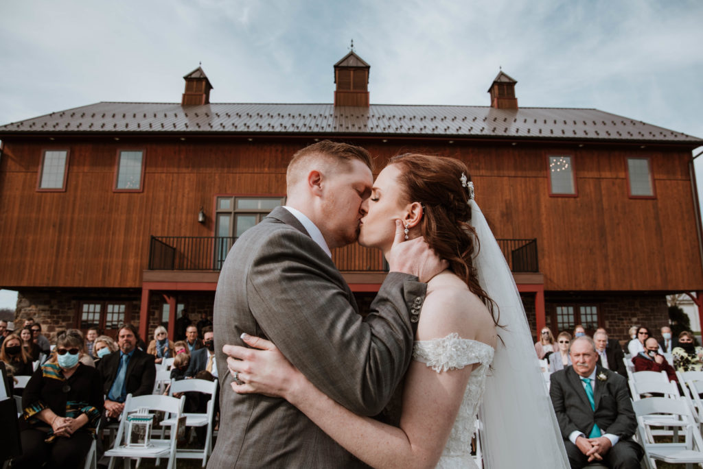 A couple is kissing in front of a farm wedding venue during their ceremony.
